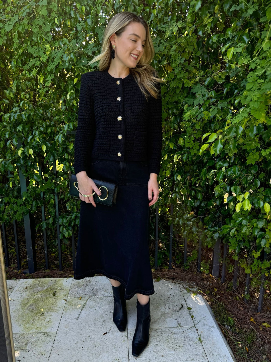 Ba&sh Gaspard Knit Cardigan in Noir paired with Volentia Denim Midi Skirt in Blackstone, Lizzie Fortunato Midnight Masquerade Earrings, Alexis Bittar Twisted Gold Side Handle Clutch Bag in Black available at Barbara Katz